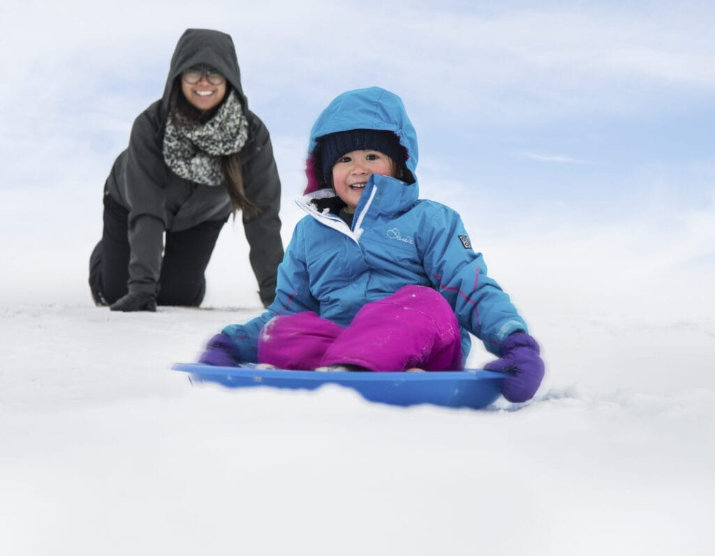 A child bundled up in a blue winter jacket and pink snow pants sledding down a snowy hill with a joyful expression, while an adult smiles in the background.
