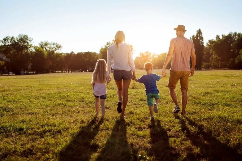 A family of four walking hand-in-hand across a grassy field at sunset. The parents hold hands with their two young children, creating a warm and peaceful moment with the sun setting in the background.