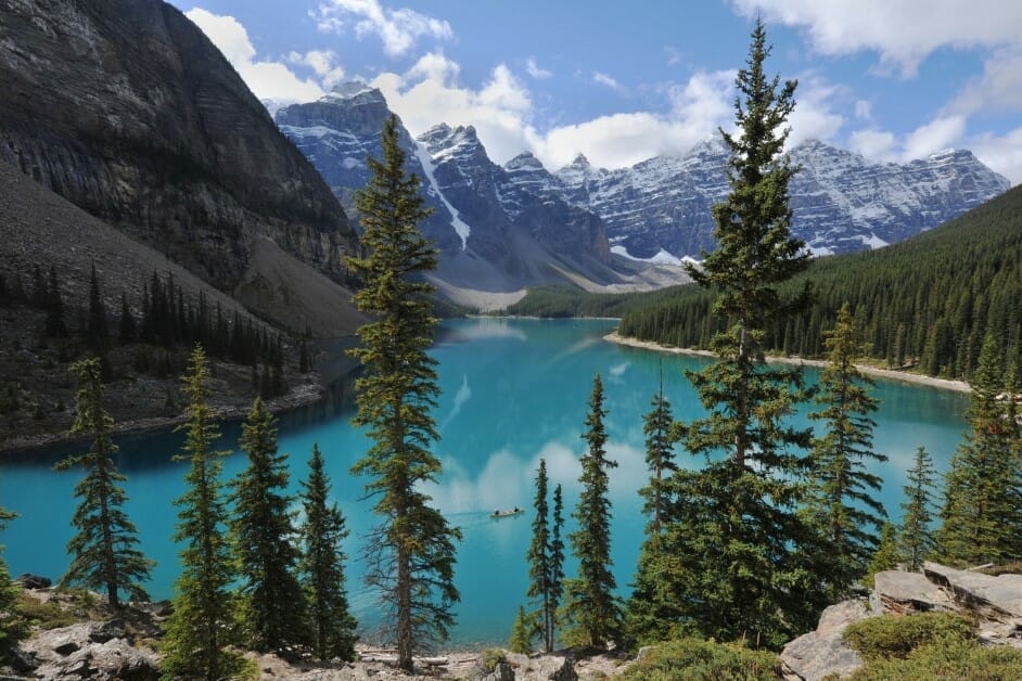 A scenic view of tall evergreen trees and dramatic snow-capped mountains. The vibrant turquoise water reflects the surrounding peaks under a partly cloudy sky.
