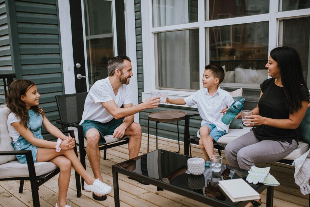 A family of four sitting together on a backyard deck, smiling and sharing drinks. The father and son clink their glasses while the mother and daughter enjoy the moment on a sunny day