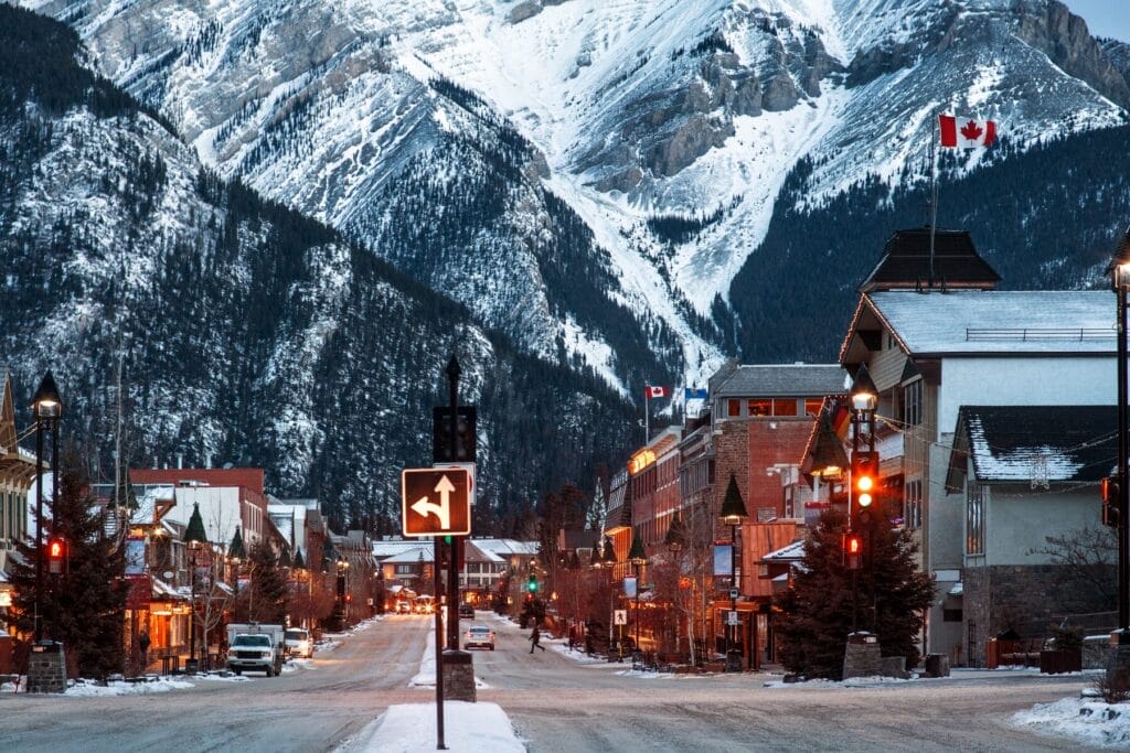 A snowy street in Banff, lined with charming buildings and lit streetlights, with majestic snow-covered mountains rising in the background and a Canadian flag waving atop a building.