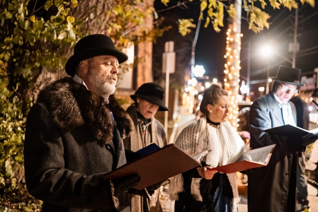 A group of carolers dressed in traditional Victorian-style clothing singing on a street at night. The street is adorned with festive lights and decorated trees