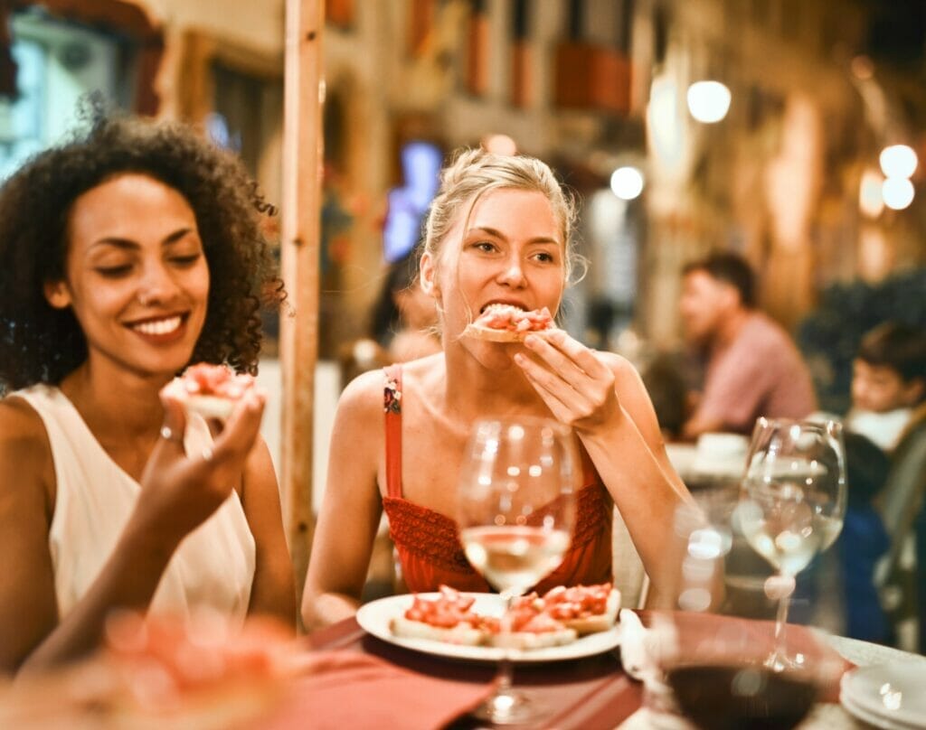Two women enjoying a meal at an outdoor restaurant in a lively evening setting. One woman is smiling while holding a bite of food, and the other is eating bruschetta. Glasses of wine are on the table, and soft lights create a warm ambiance.