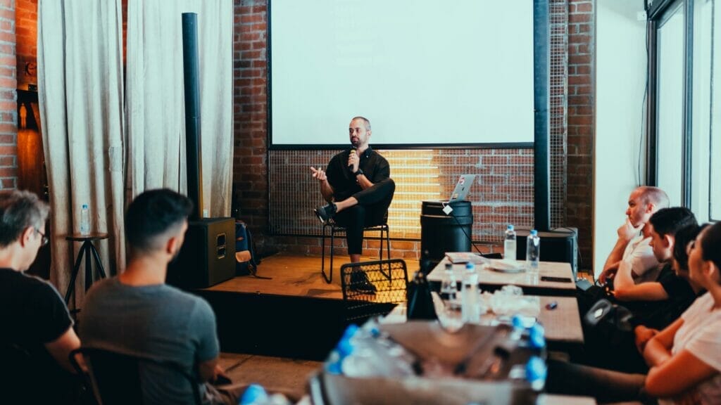 A speaker sits on a stool on stage, holding a microphone and engaging with an audience in a modern, industrial-style venue. Attendees sit at tables, listening attentively, with a presentation screen in the background.