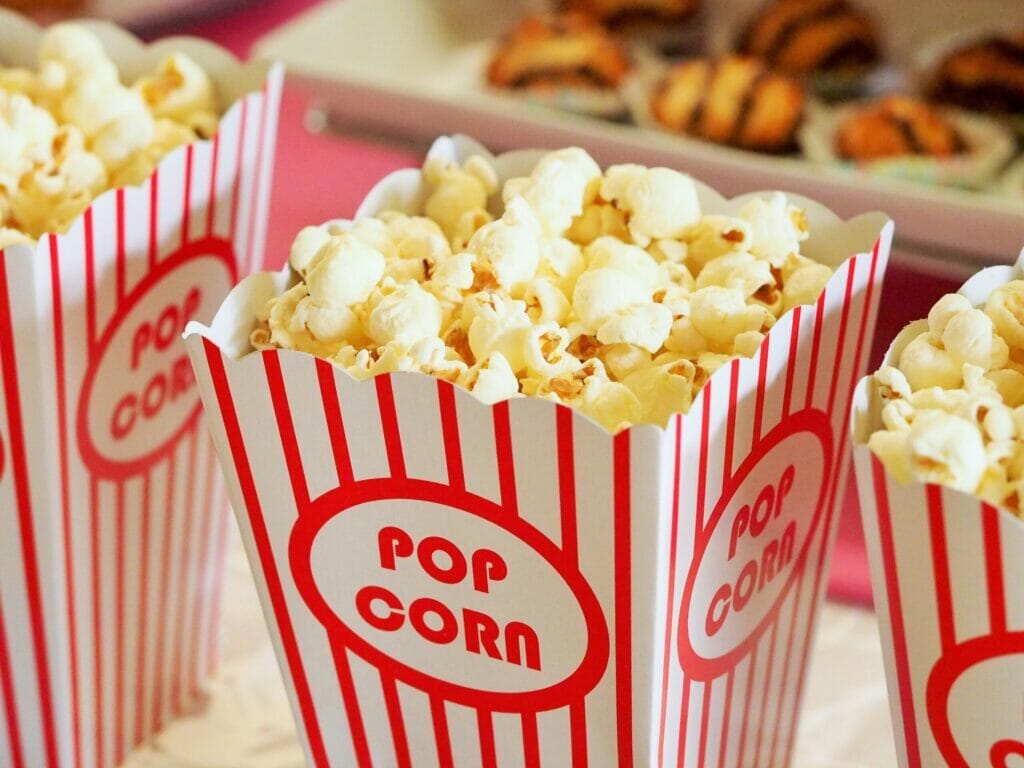 Three red-and-white striped popcorn boxes filled with freshly popped popcorn. In the background, blurred baked treats are visible on a pink table.