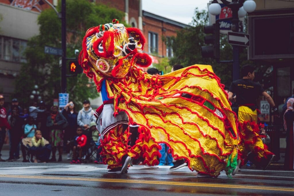 A vibrant lion dance performance during a street festival, with performers dressed in a red and yellow lion costume. The lion's head is detailed and colorful, surrounded by spectators watching from the sidewalk on a cloudy day.