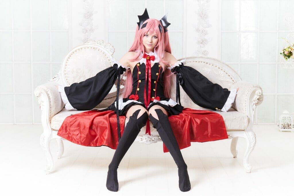 A cosplayer dressed in a gothic-inspired outfit with long pink hair, sitting on an ornate white sofa. The costume features a black and red dress with dramatic sleeves, thigh-high stockings, and decorative black horns, set against a bright, elegant background.
